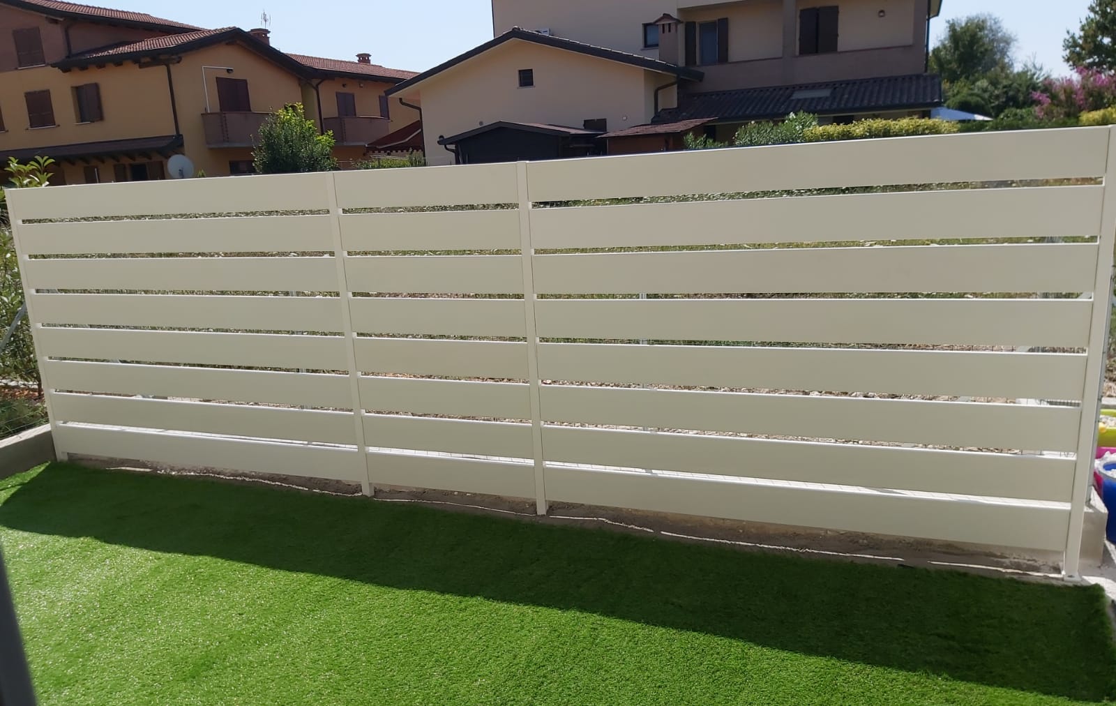 Recinzione orizzontale color crema di fronte a un prato artificiale verde, con edifici sullo sfondo in una giornata di sole.