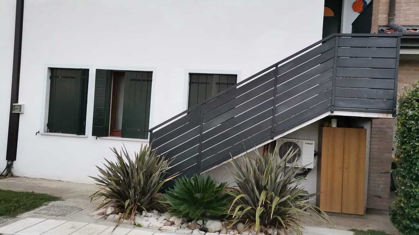 Edificio bianco con scala nera, persiane verdi e piante sulla facciata.