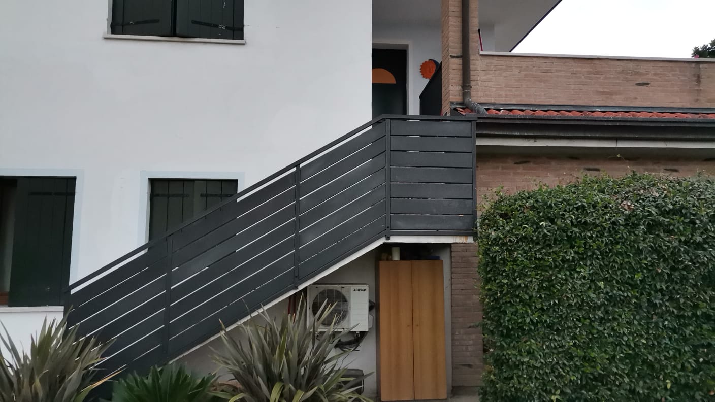 Esterno di una casa bianca con una scala e una ringhiera grigio scuro. C'è una porta di legno e vegetazione incolta.