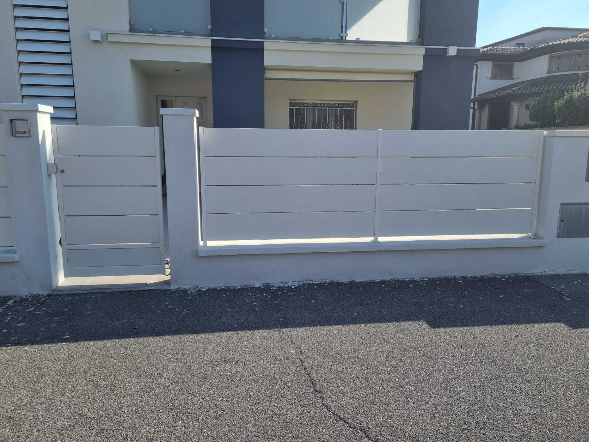 Recinzione bianca con listelli orizzontali e cancello di fronte a un edificio chiaro, situato su una strada asfaltata scura.