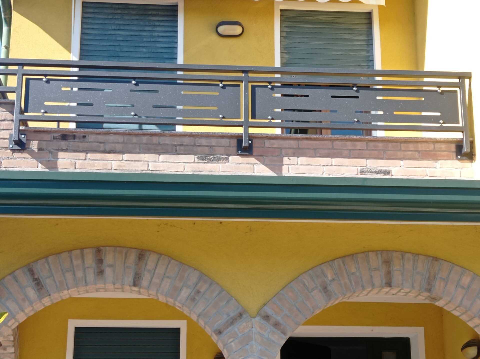Ringhiera in metallo nero di un balcone su un edificio giallo con supporti in mattoni ad arco e persiane blu.