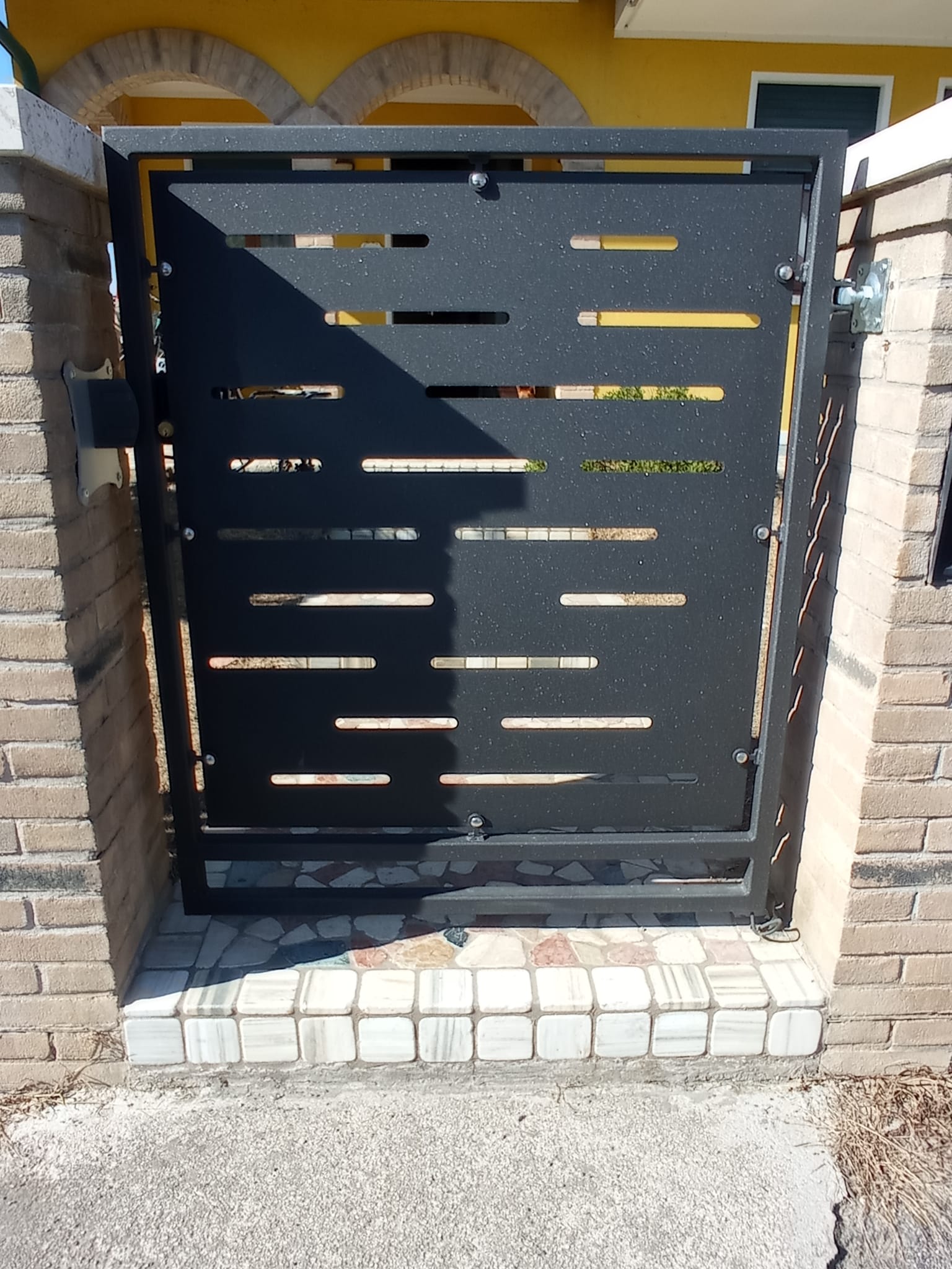 Cancello in metallo nero con ritagli orizzontali inseriti in colonne di mattoni.