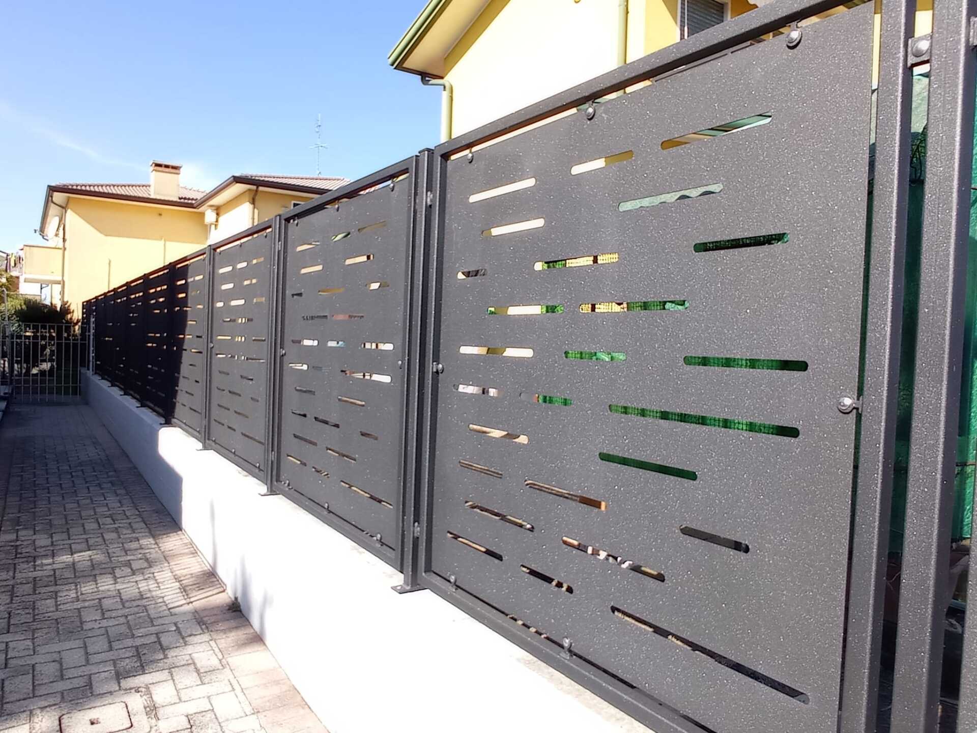 Pannelli di recinzione in metallo con motivi ritagliati orizzontali, montati su un muro bianco, lungo un marciapiede.