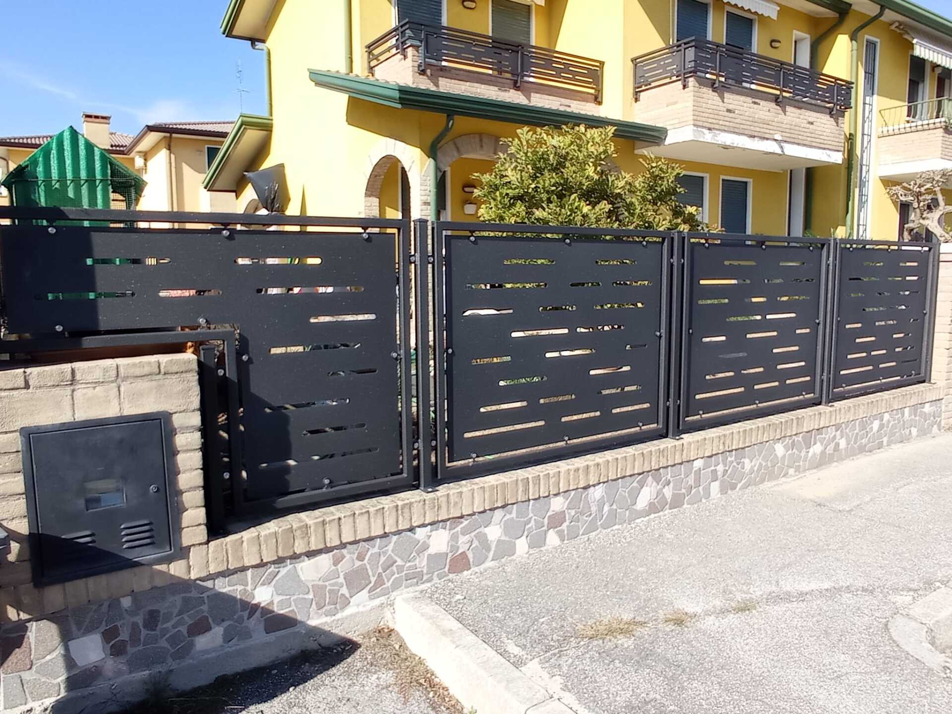 Recinzione in metallo nero con ritagli orizzontali sopra un muro di mattoni e pietra, di fronte a un edificio giallo.