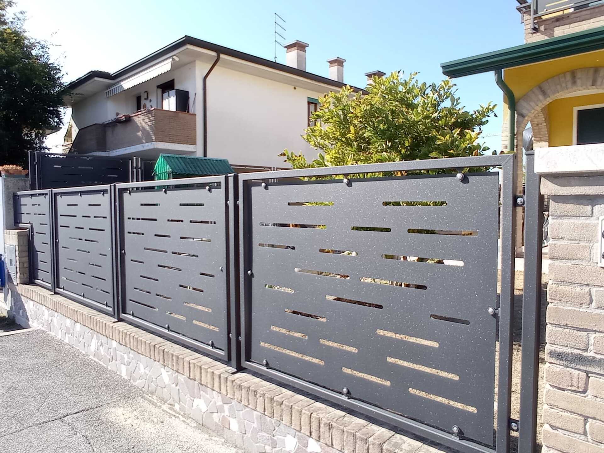 Recinzione in metallo grigio con ritagli orizzontali sopra un muro di mattoni di fronte a una casa.