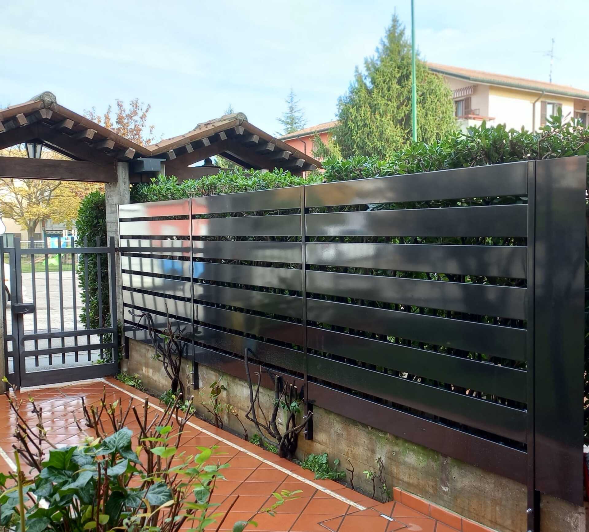 Recinzione in metallo nero a doghe orizzontali accanto a un patio piastrellato e un cancello, con siepi verdi.