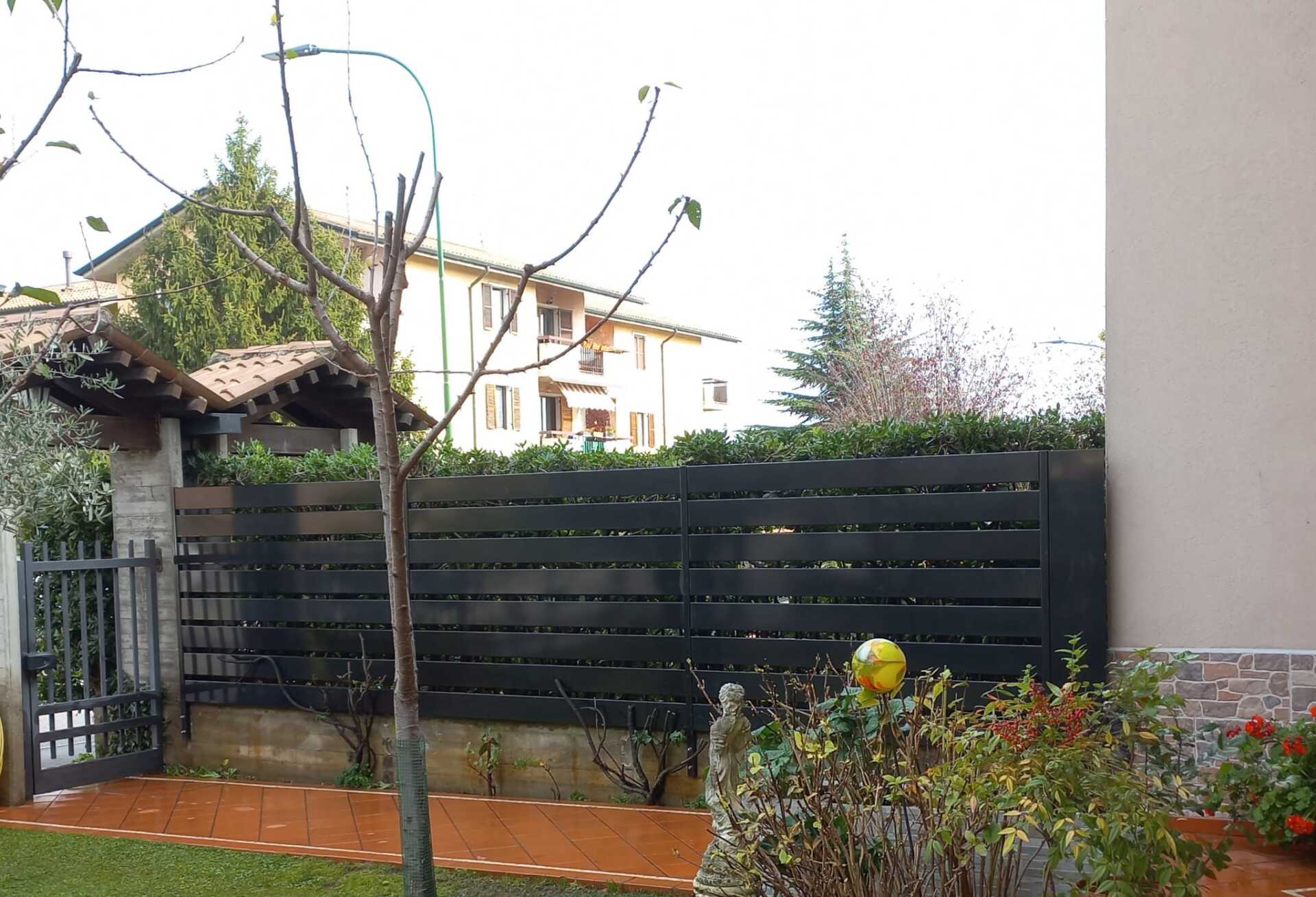 Recinzione in legno nero con cancello davanti a una casa. Albero spoglio in primo piano, cespugli ed erba.