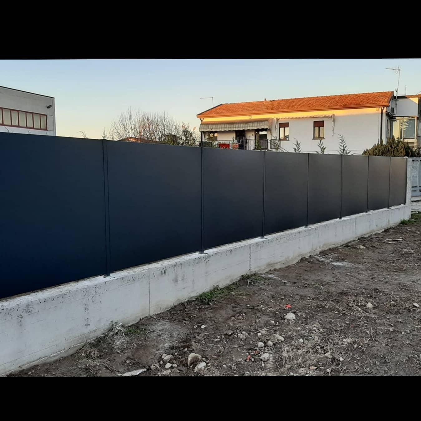 Recinzione in metallo grigio scuro su una base di cemento, al confine con un cantiere edile. Case sullo sfondo.