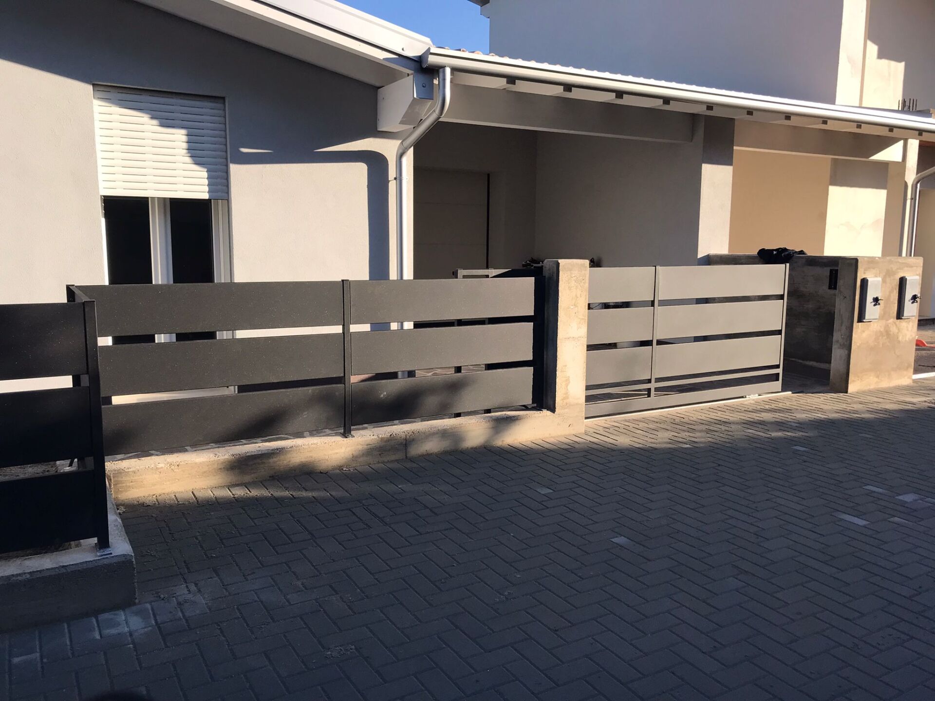 Recinzione in metallo nero con doghe orizzontali davanti a una casa moderna, vialetto pavimentato.