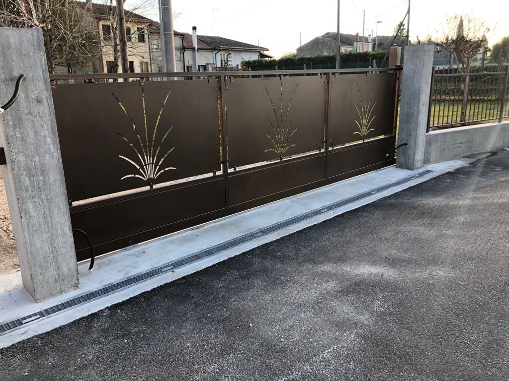 Cancello in metallo marrone con ritagli decorativi, inserito tra pilastri di cemento e un vialetto pavimentato.