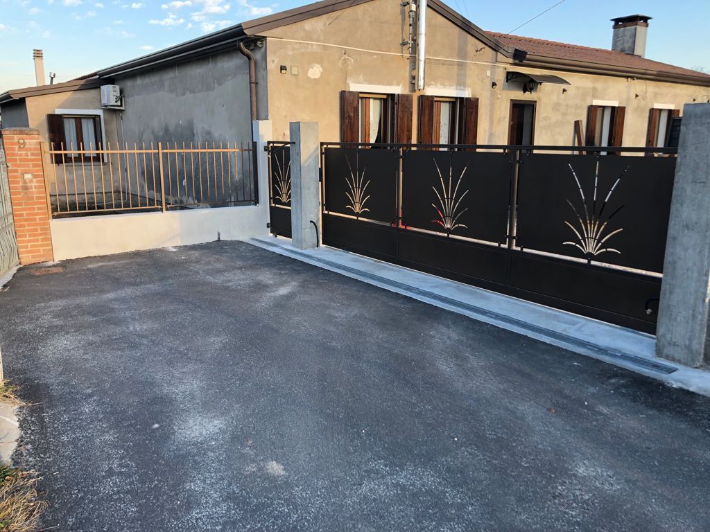 Cancello nero con motivo decorativo davanti a una casa in cemento con persiane marroni; vialetto d'accesso.