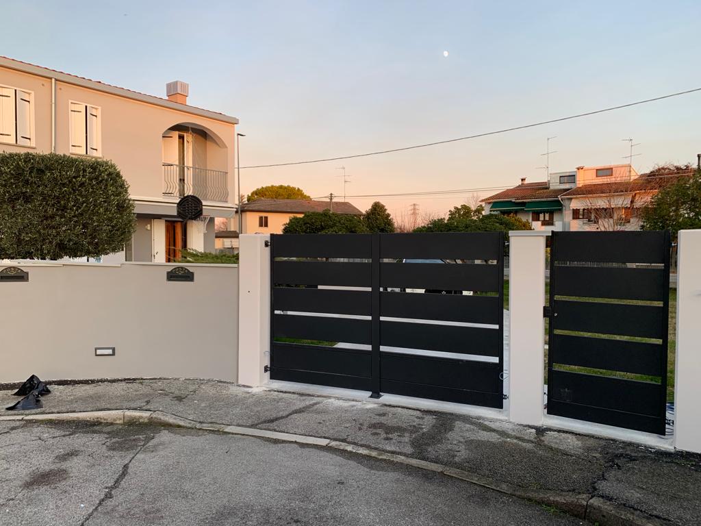 Un moderno cancello e una recinzione in metallo nero davanti a una casa chiara al tramonto.