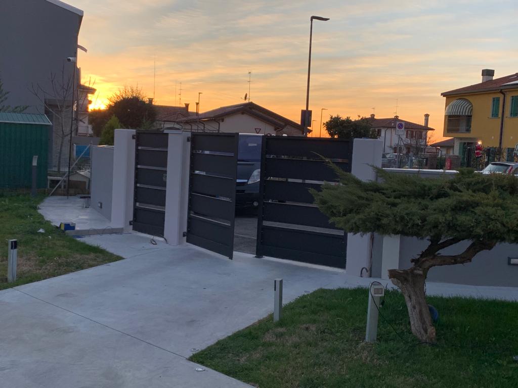 Cancello nero che si apre su un vialetto al tramonto, con edifici sullo sfondo.