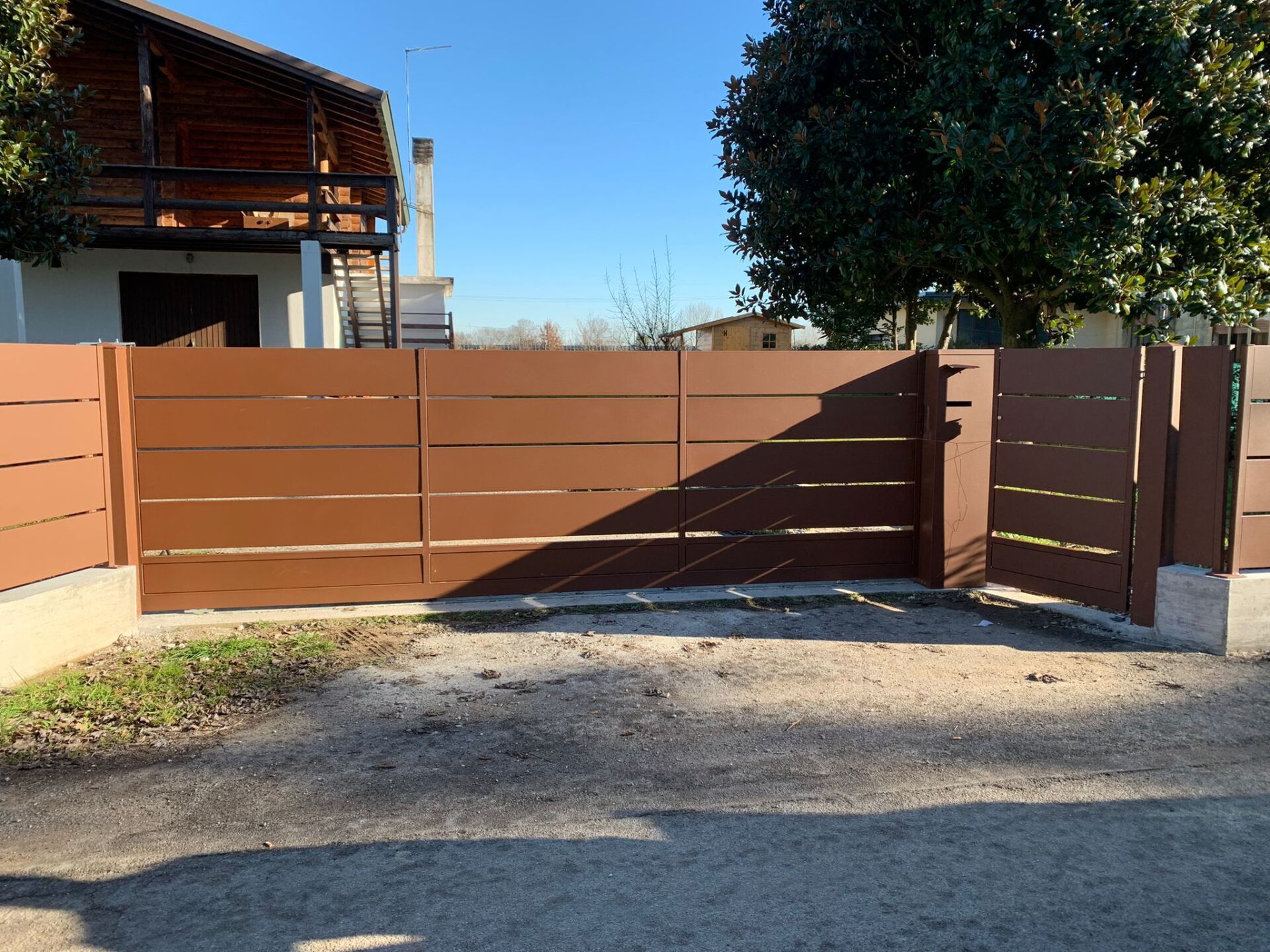 Cancello marrone a listelli orizzontali davanti a una casa, appoggiato su cemento e asfalto. Giornata di sole.