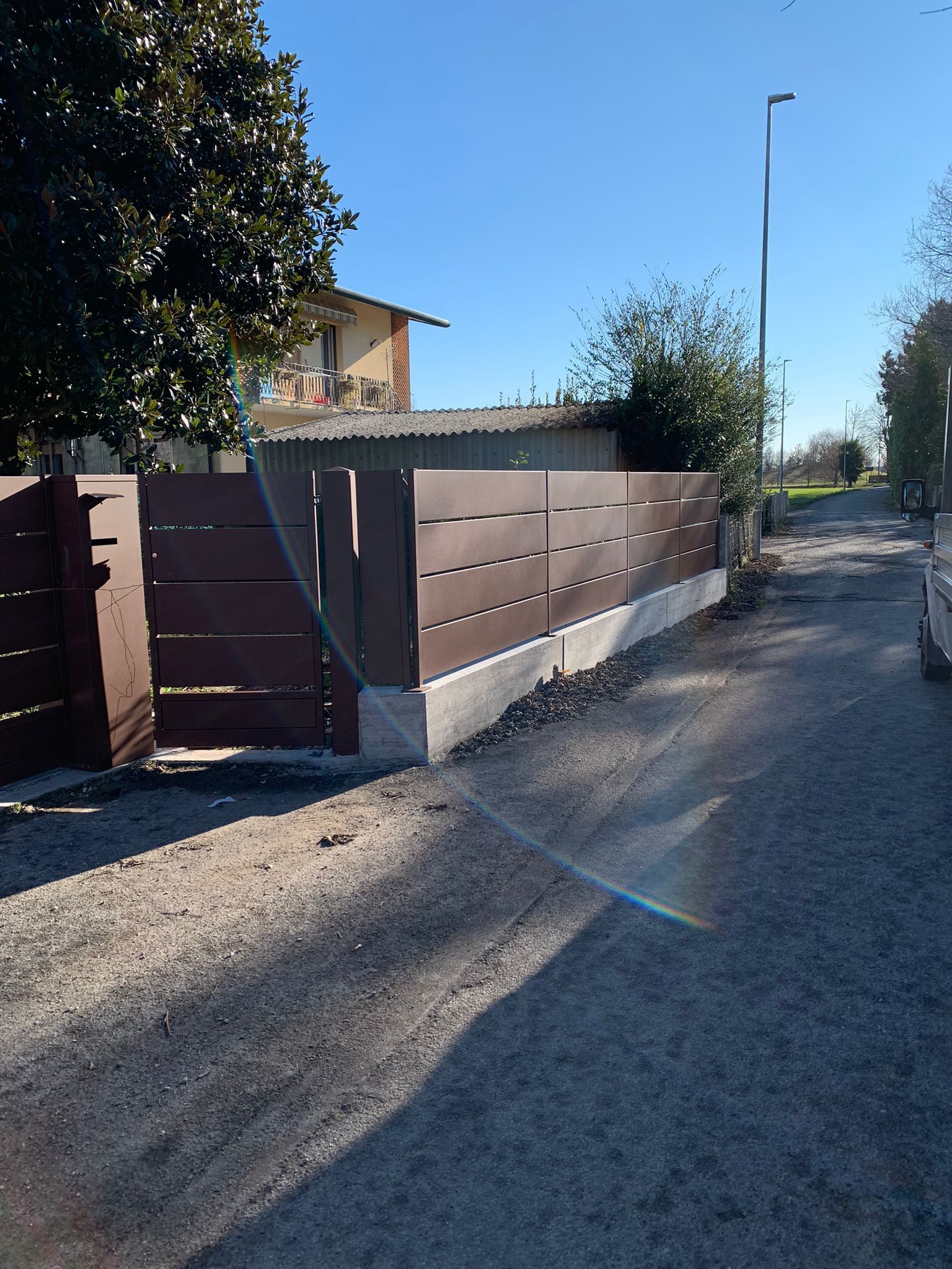 Recinzione marrone con listelli orizzontali, cancello, base in cemento e vialetto in ghiaia.