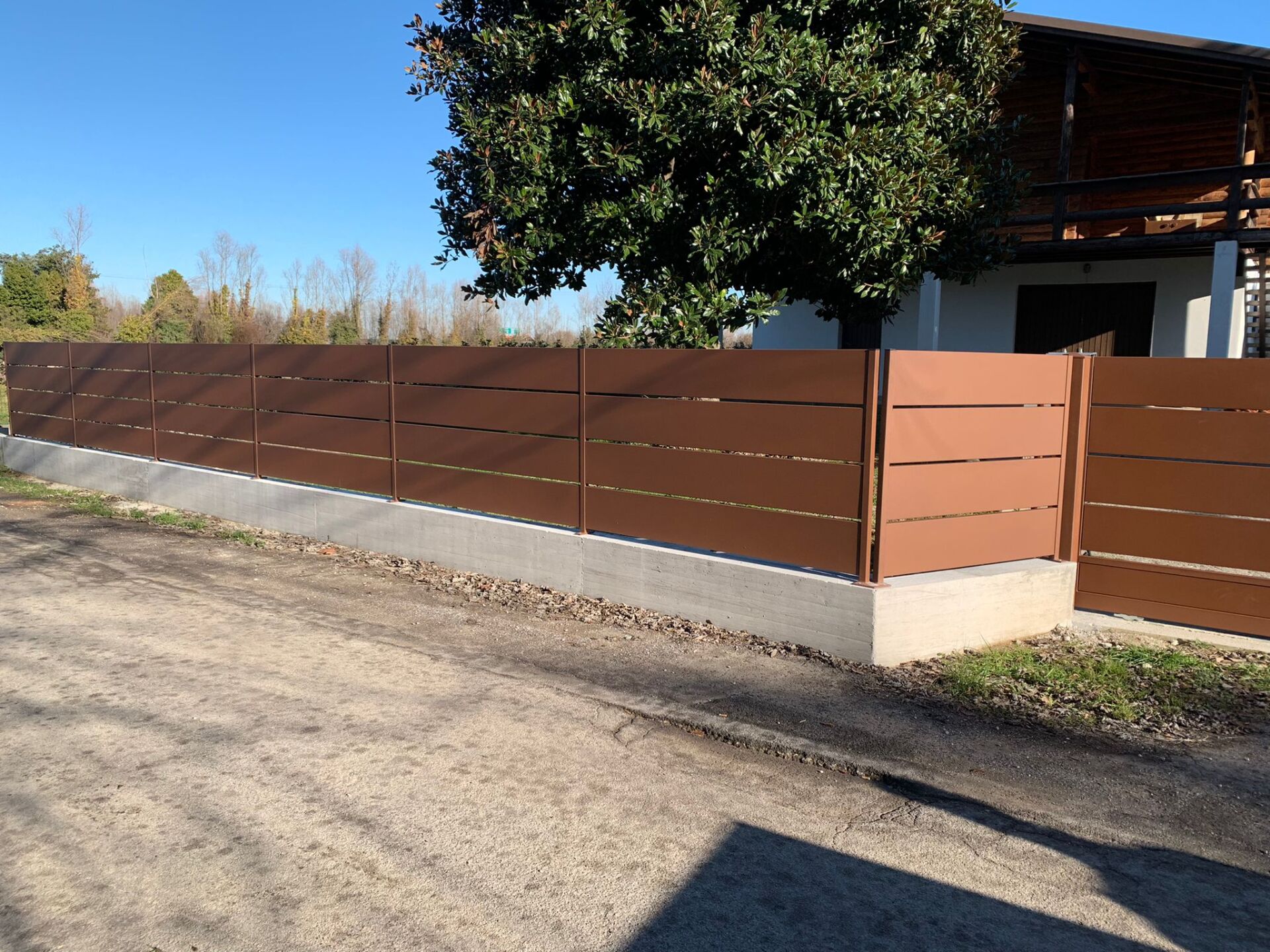 Recinzione in legno marrone su una base di cemento lungo una strada sterrata, con una casa e un albero sullo sfondo.