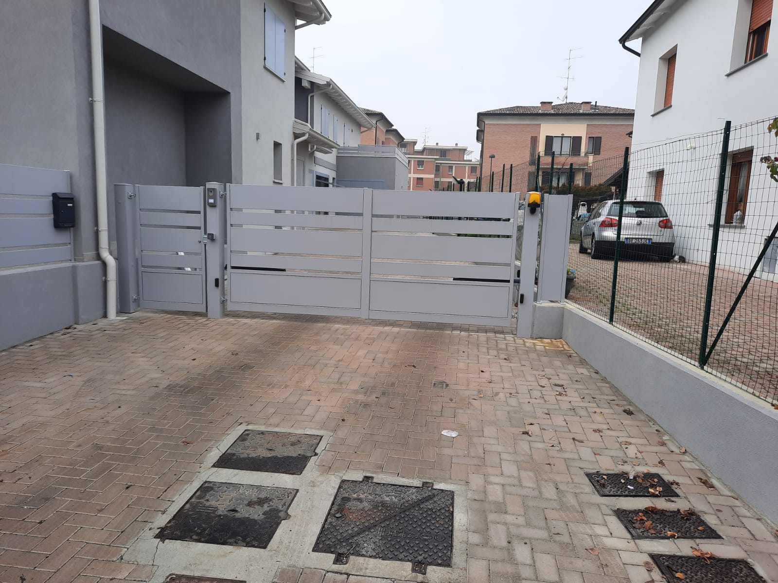 Cancello grigio con stecche orizzontali che conduce a una casa. Vialetto in mattoni, altre case sullo sfondo.