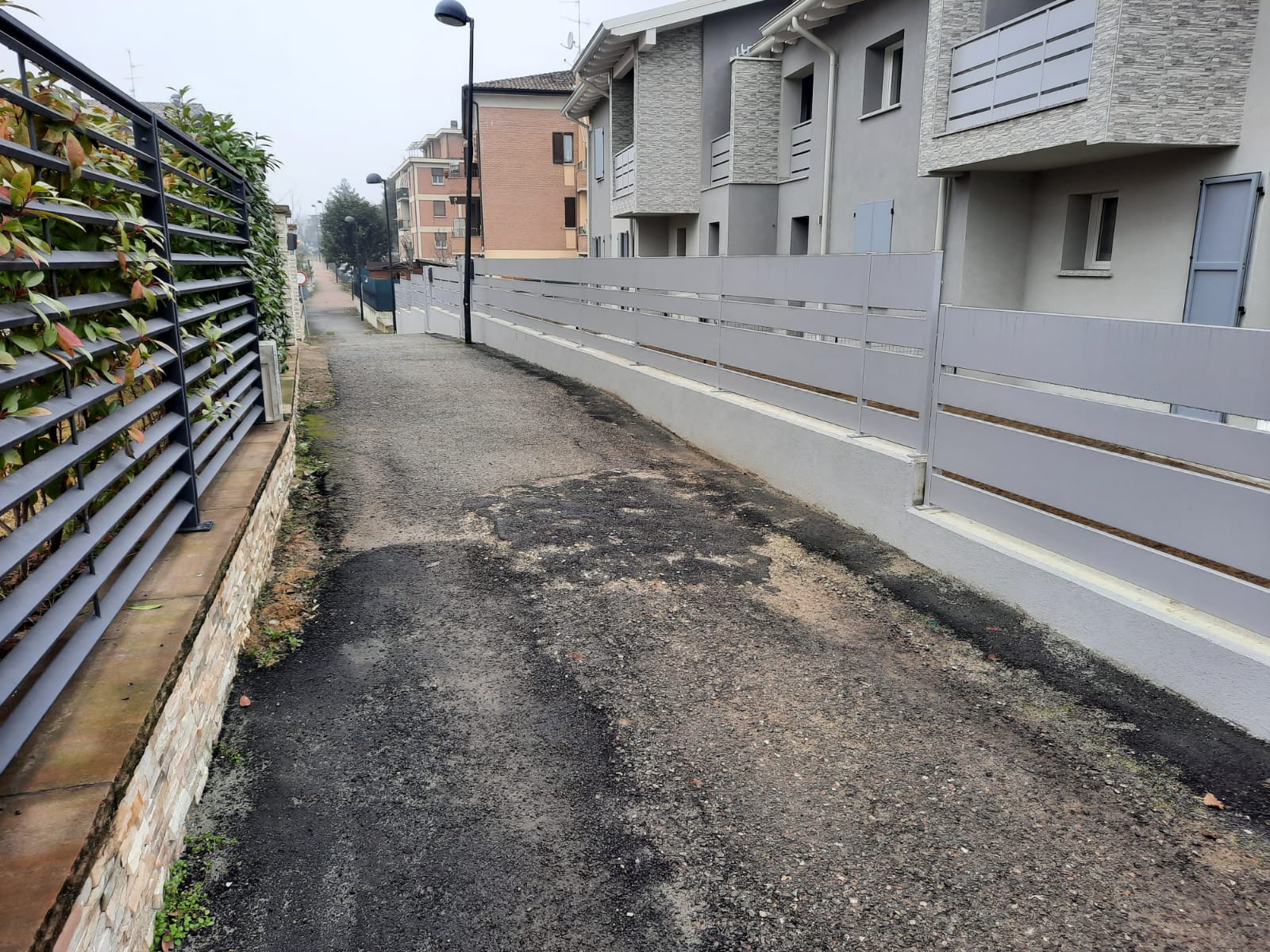 Stretto sentiero asfaltato tra una recinzione metallica con fogliame e una recinzione di legno grigio accanto agli edifici.