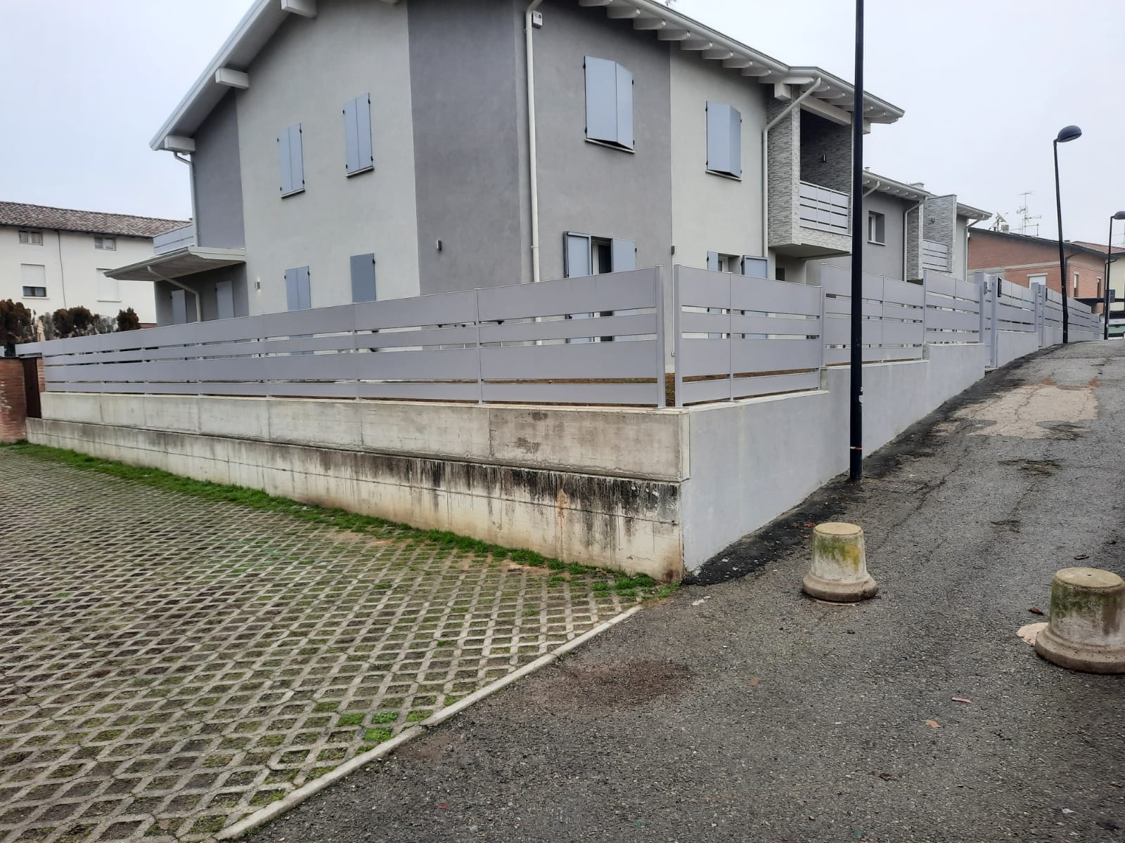 Casa grigia a due piani dietro una recinzione grigia in cima a un muro di cemento lungo una strada asfaltata.