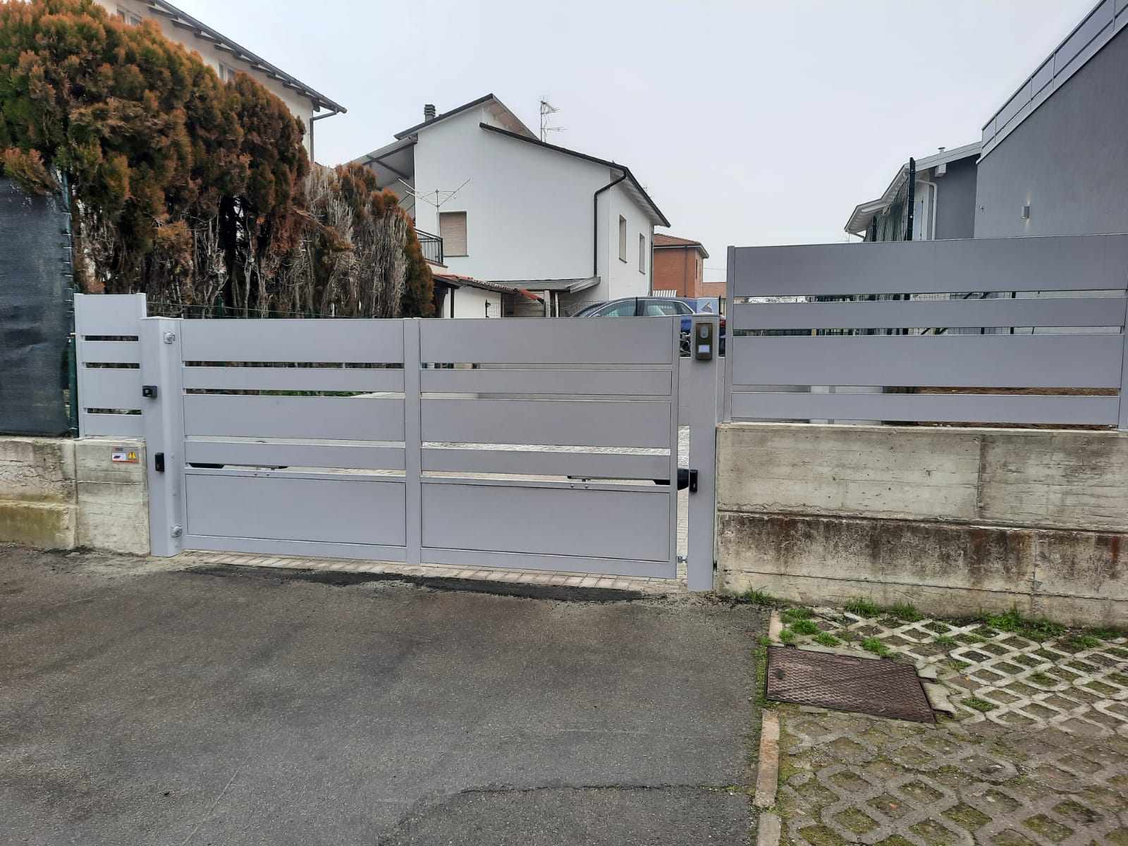 Cancello e recinzione in metallo grigio davanti a una casa. Il cancello è chiuso da un muro di cemento.