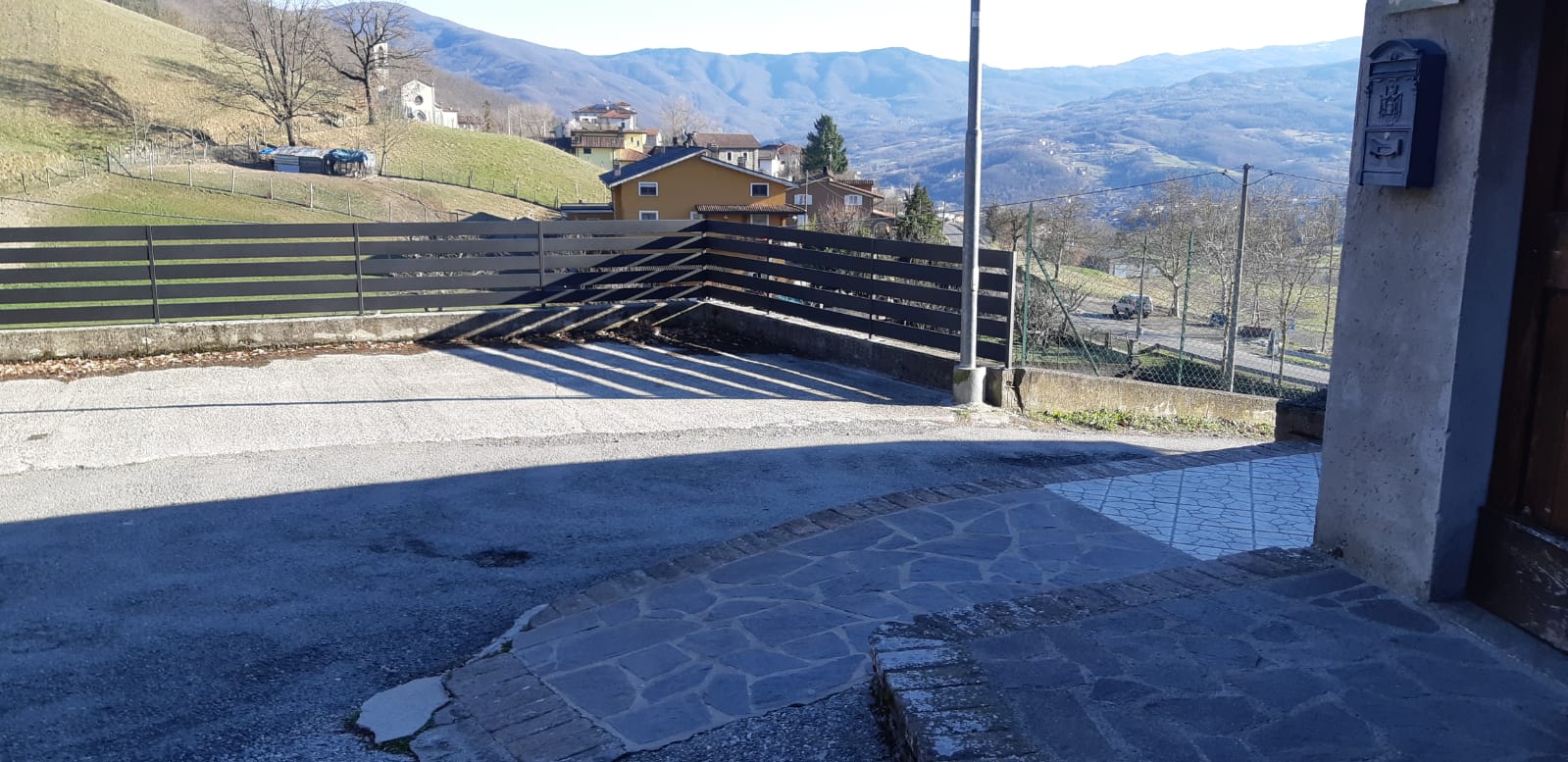 Vista di un paesaggio montano da un'area pavimentata, con una recinzione e il muro di un edificio sulla destra.