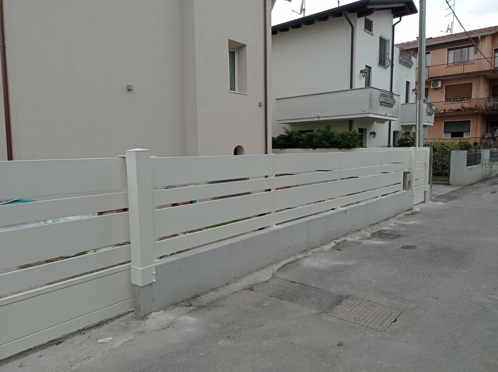 Recinzione bianca a doghe orizzontali lungo un muro grigio, accanto a una strada con edifici sullo sfondo.