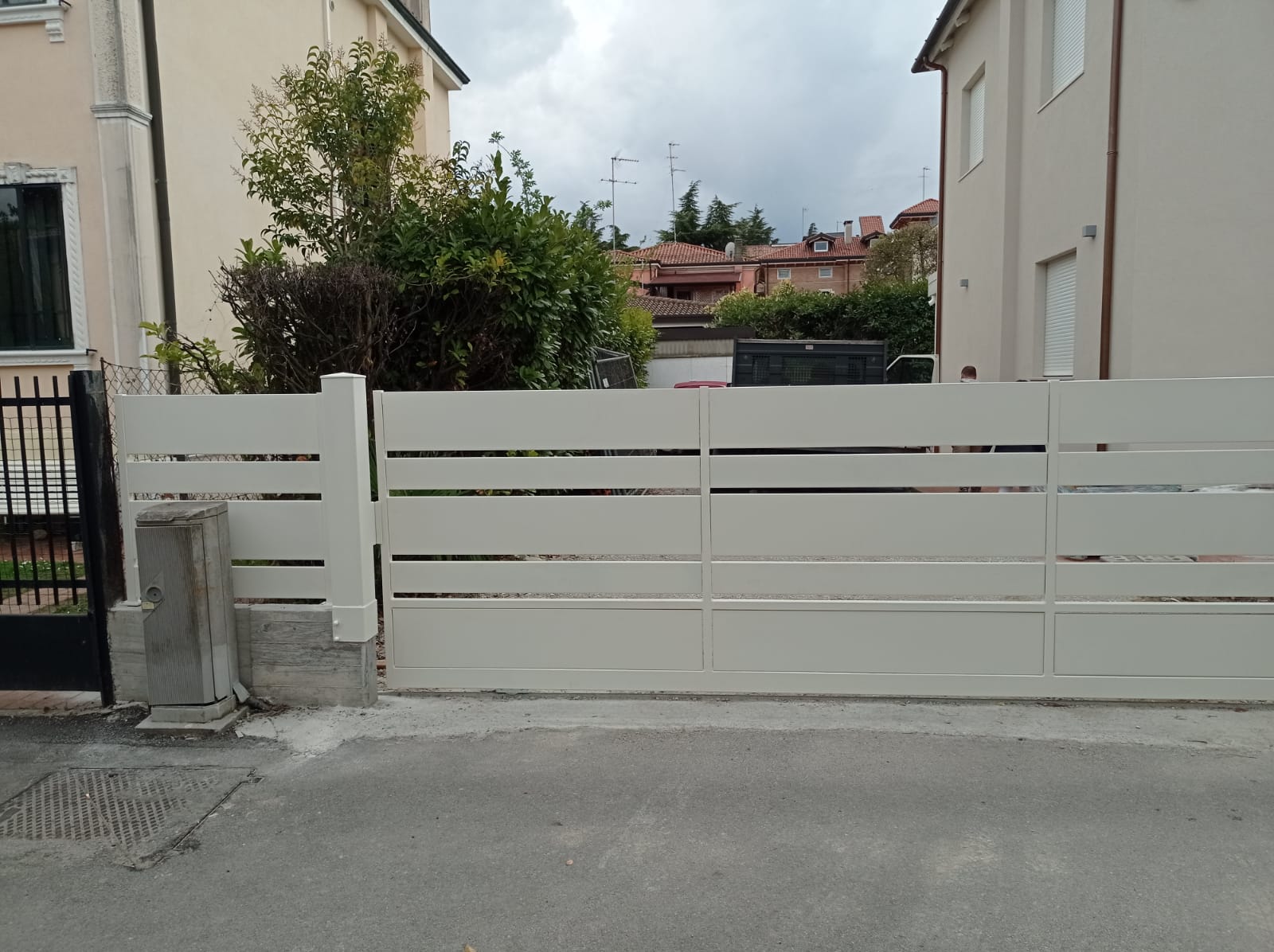 Cancello bianco a stecche orizzontali con una piccola scatola grigia sul lato, che blocca un vialetto. Case sullo sfondo.