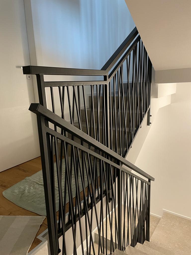 Scala con ringhiera in metallo nero con barre verticali ondulate; pareti e moquette di colore chiaro.