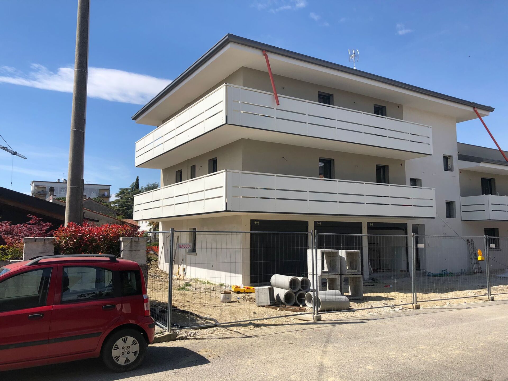 Edificio moderno di tre piani in costruzione con balconi bianchi e un'auto rossa parcheggiata di fronte.
