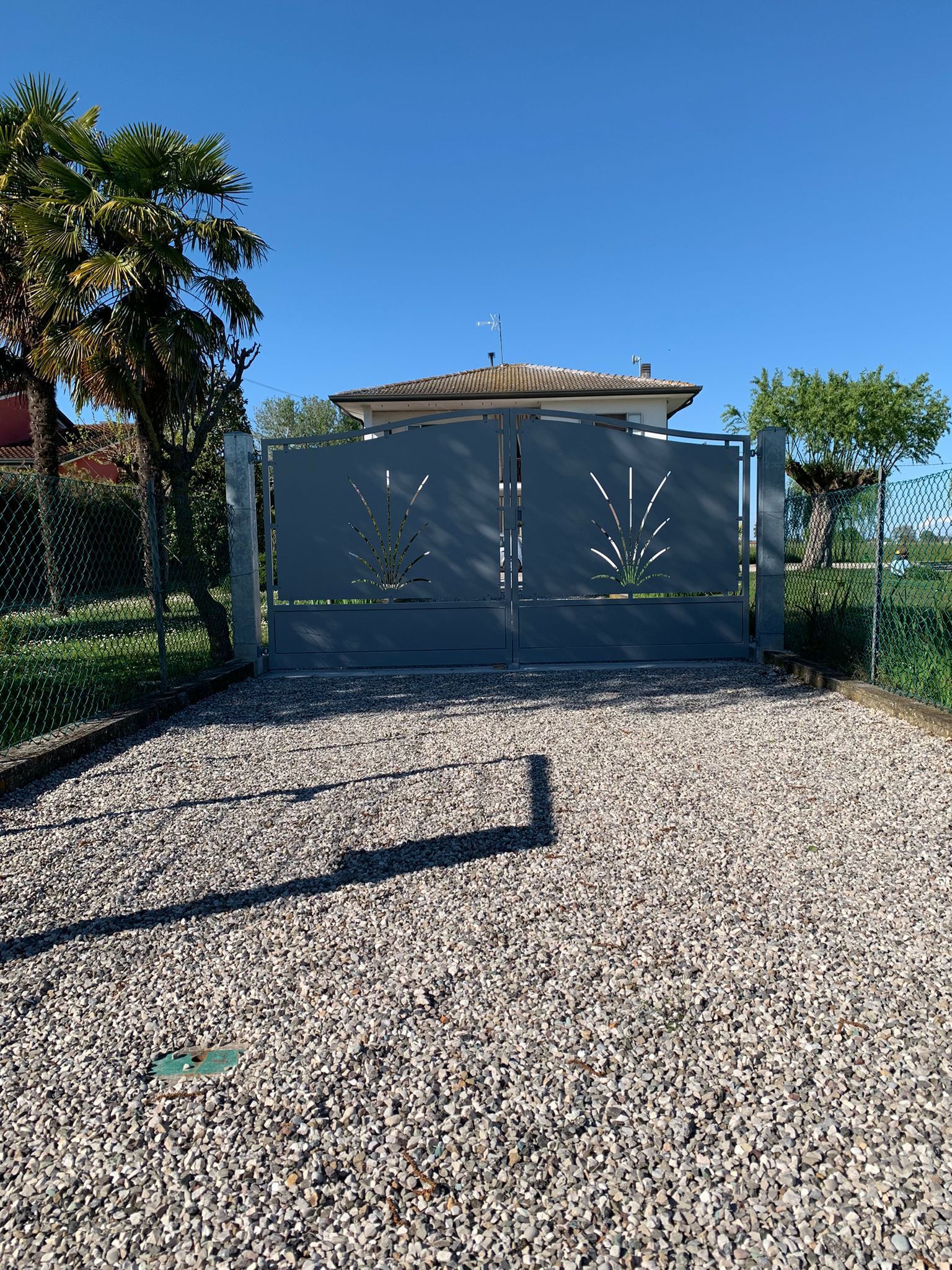 Cancelli in metallo grigio con ritagli decorativi, vialetto di ghiaia, casa sullo sfondo, cielo sereno.