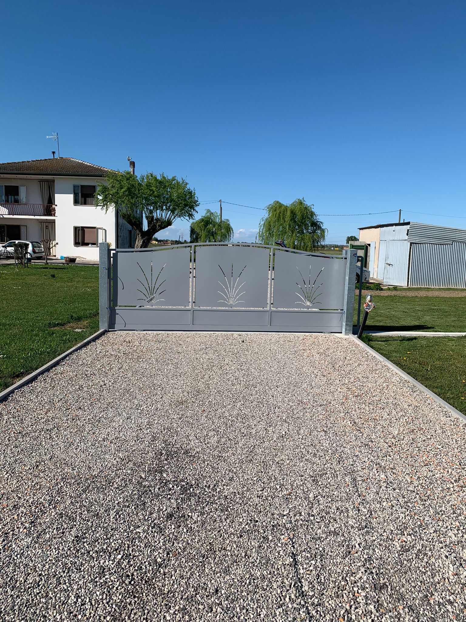 Cancello di metallo grigio su un vialetto di ghiaia, con una casa e alberi sullo sfondo sotto un cielo azzurro.