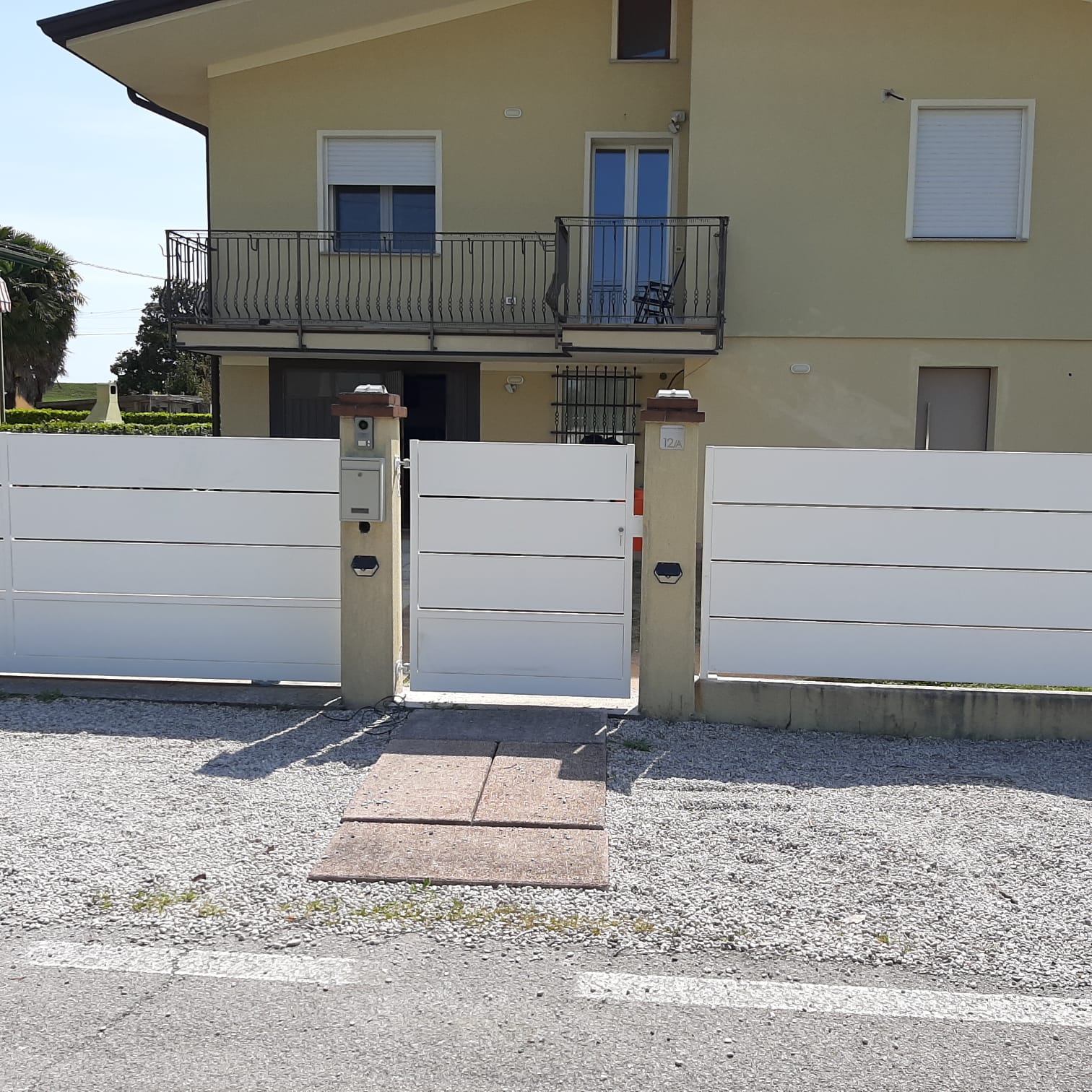 Cancello bianco e recinzione davanti a una casa gialla con balcone e finestre. Un sentiero in pietra conduce al cancello.