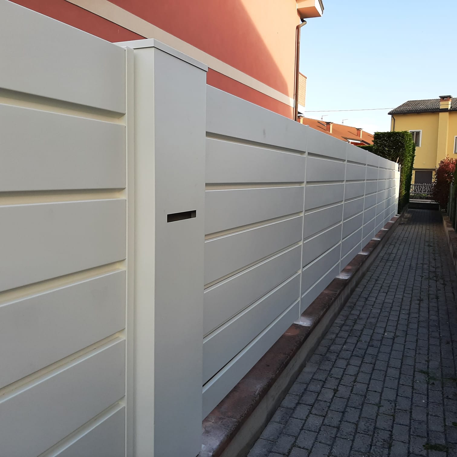 Recinzione bianca con listelli orizzontali lungo un vialetto in mattoni accanto a un edificio.