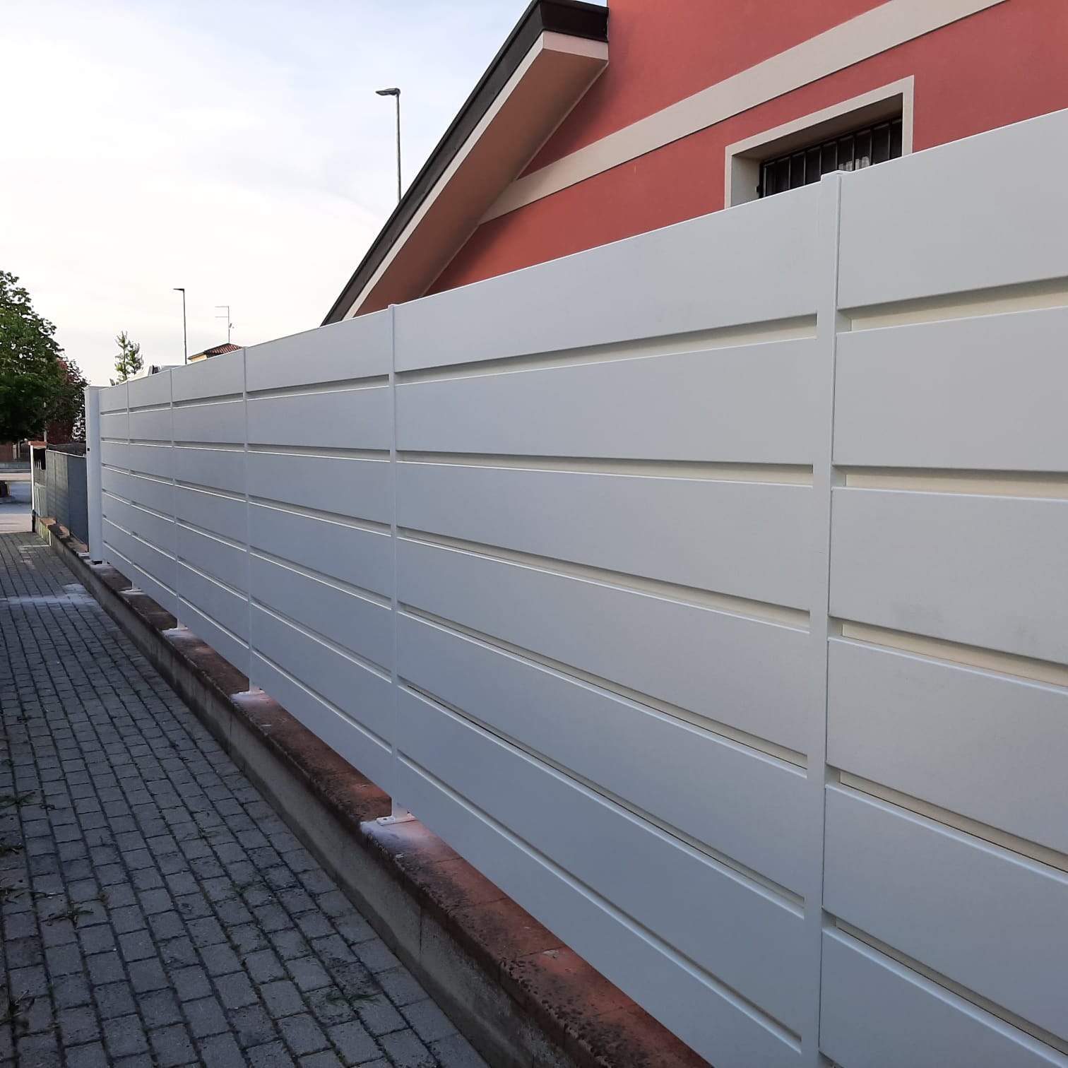Recinzione bianca con listelli orizzontali lungo un marciapiede in mattoni, con un edificio rosso sullo sfondo.
