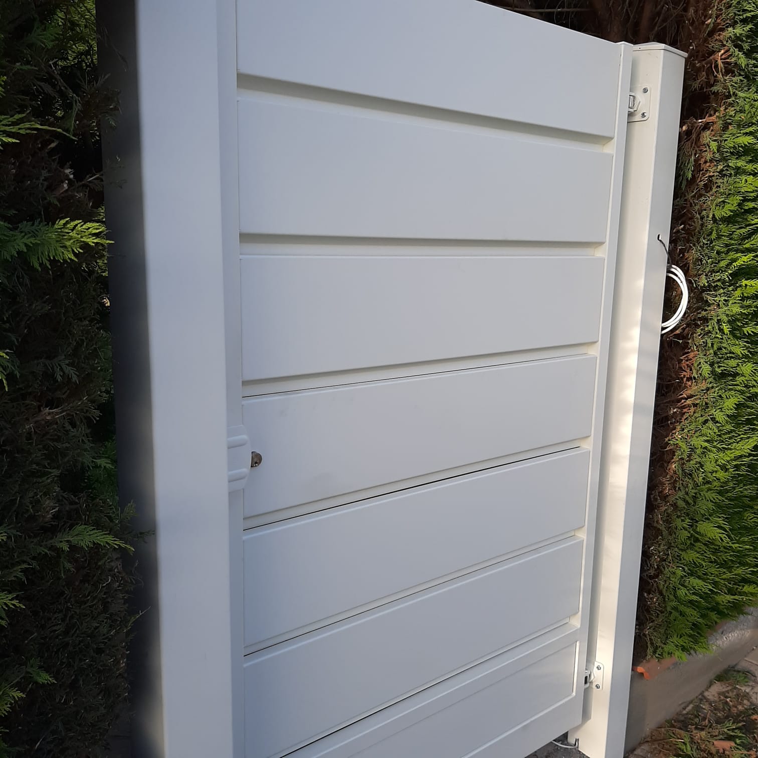 Cancello metallico bianco a stecche orizzontali, parzialmente aperto, con maniglia, di fronte a una siepe.