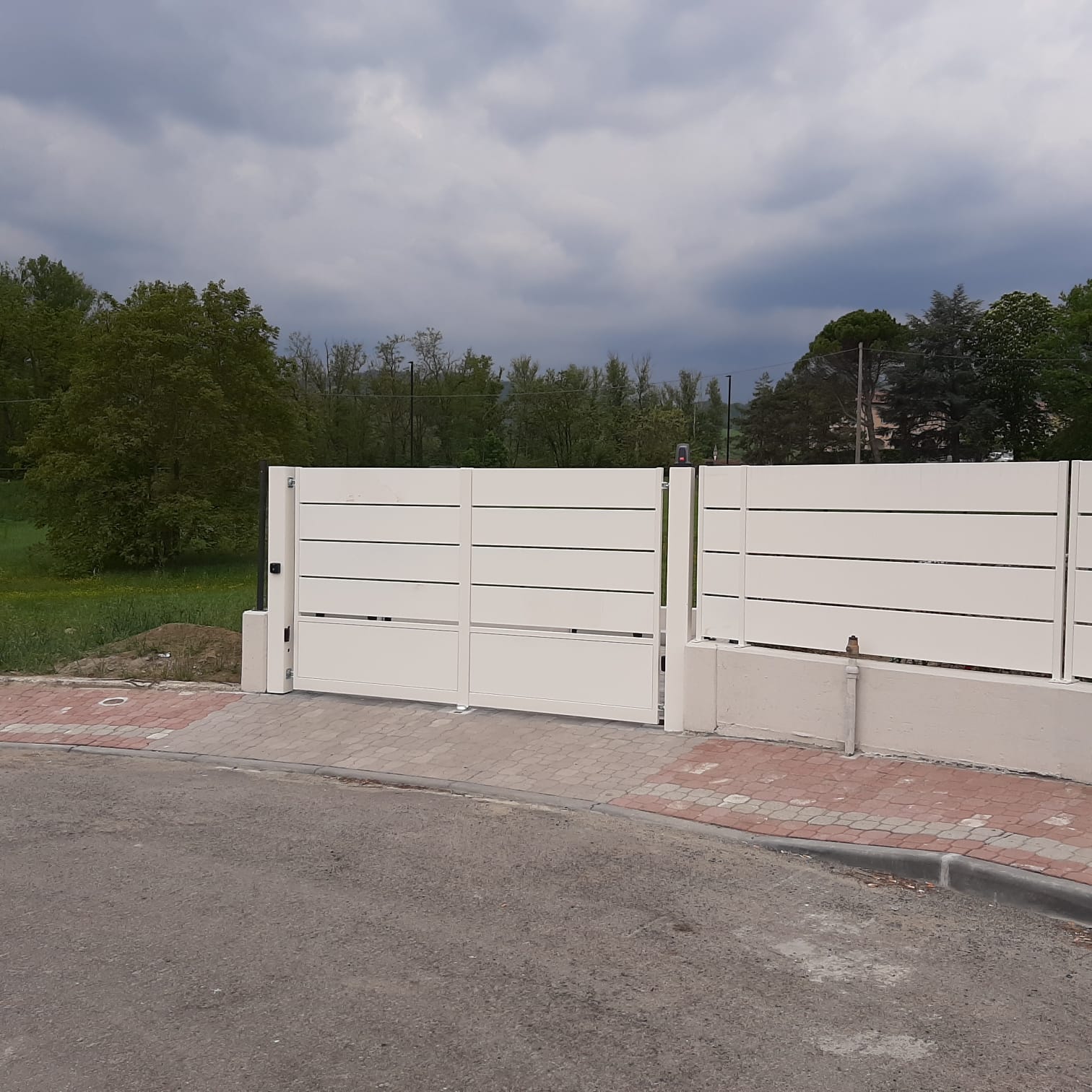 Cancello e recinzione bianchi a doghe orizzontali su supporti in cemento. Strada asfaltata e fogliame verde sullo sfondo.