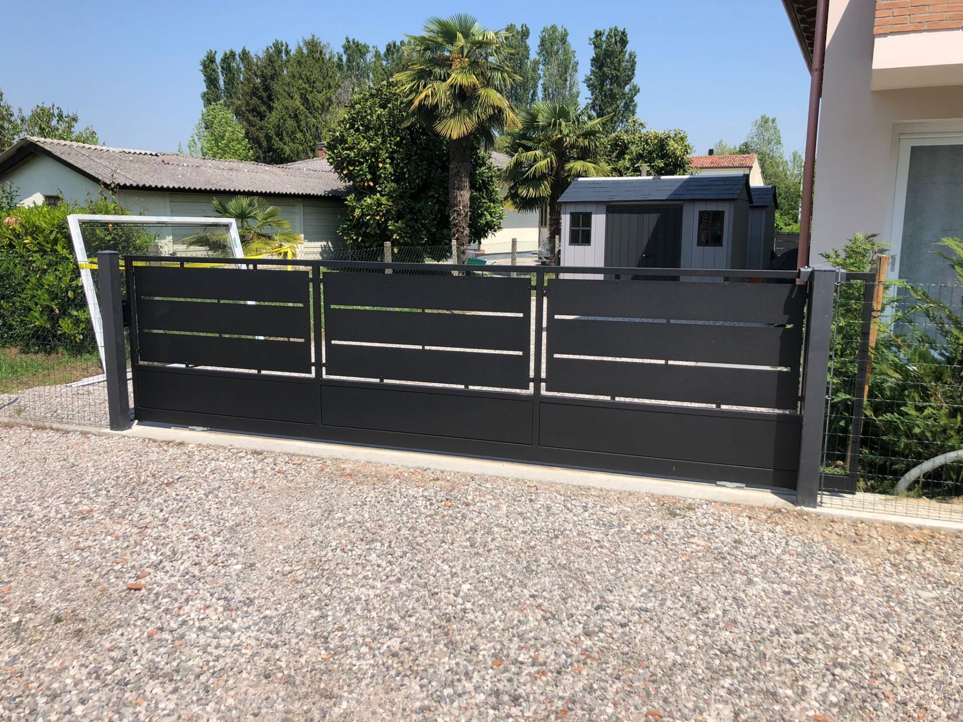 Cancello scorrevole in metallo grigio scuro con stecche orizzontali, vialetto in ghiaia.