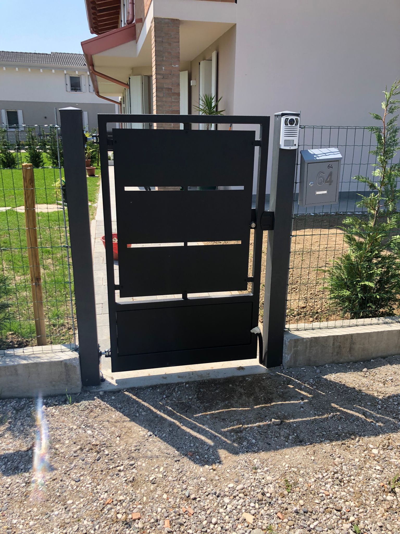 Cancello in metallo nero con ritagli orizzontali davanti a una casa, terreno ghiaioso e cassetta della posta.