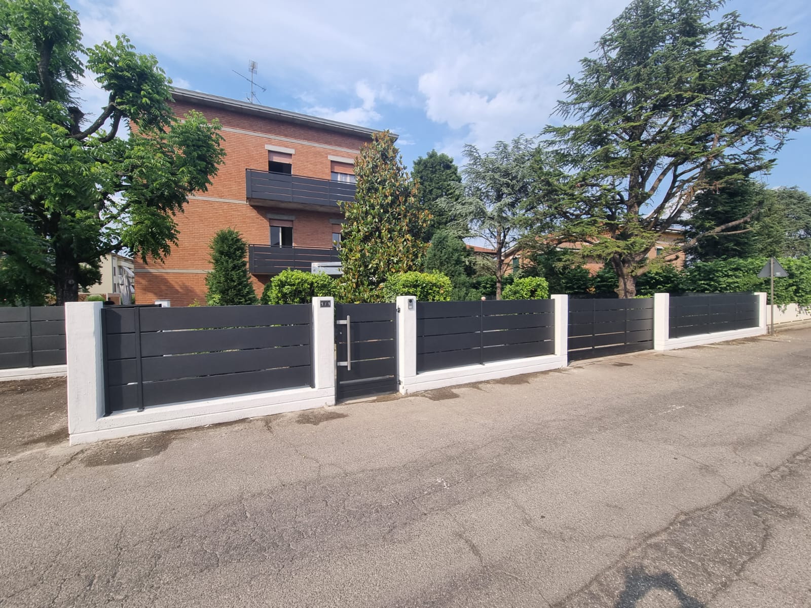 Recinzione nera a doghe orizzontali con pilastri bianchi e cancello di fronte a un edificio in mattoni.