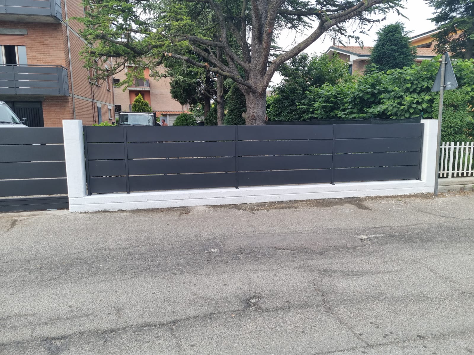 Recinzione orizzontale grigio scuro con rifiniture bianche tra gli edifici e la strada.