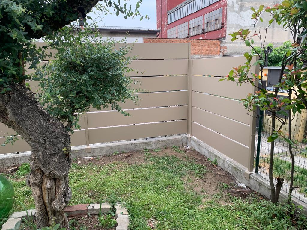 Recinzione orizzontale in doghe color marrone chiaro in un cortile, con un albero sulla sinistra e un po' di erba.