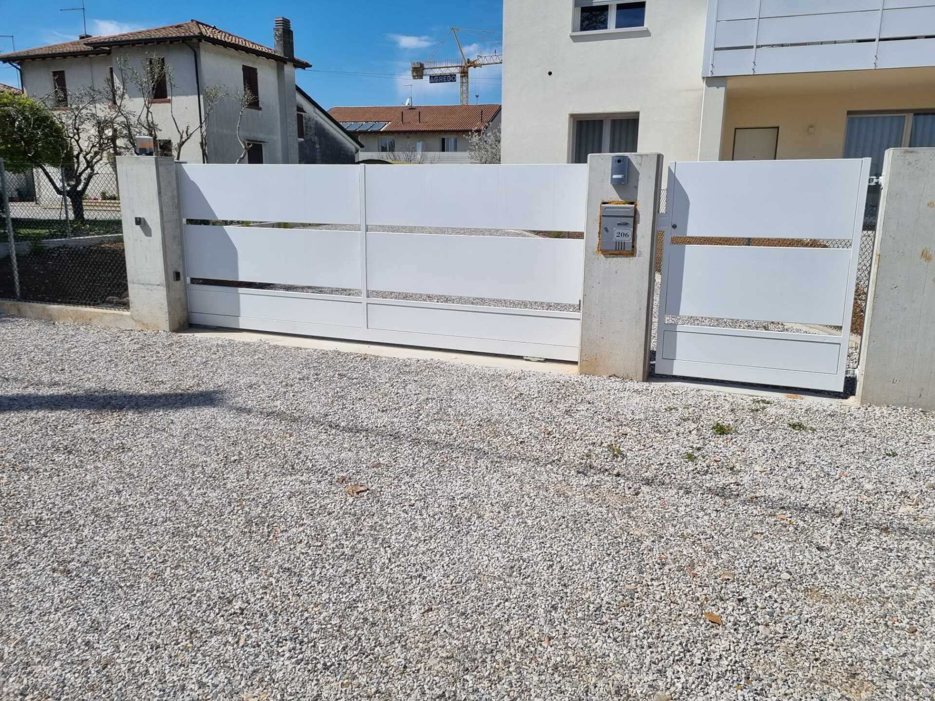 Cancello scorrevole bianco con ritagli orizzontali, cassetta postale e vialetto di ghiaia.