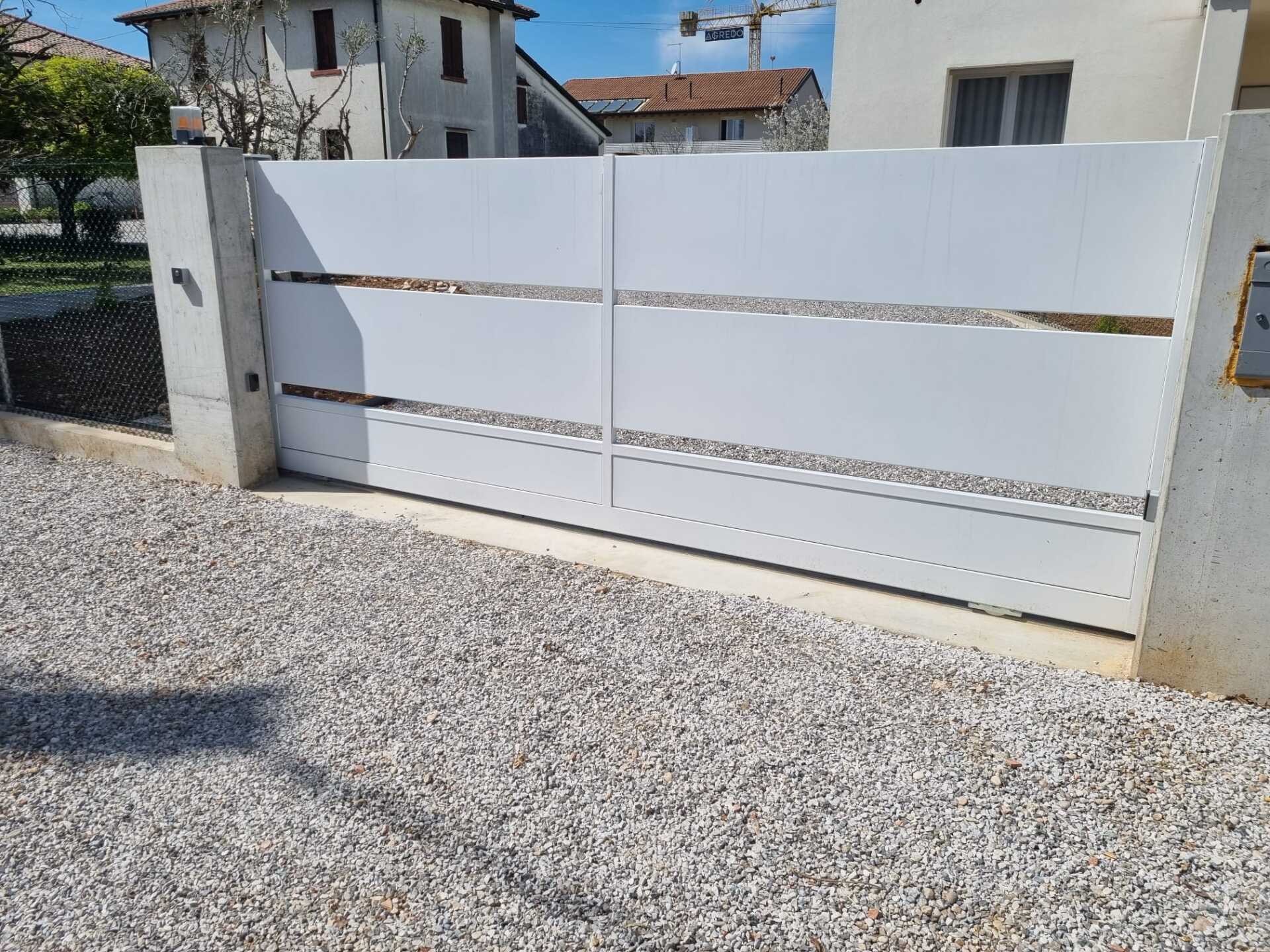 Cancello carrabile in metallo bianco con ritagli orizzontali, inserito in un vialetto di ghiaia, di fronte a una casa.