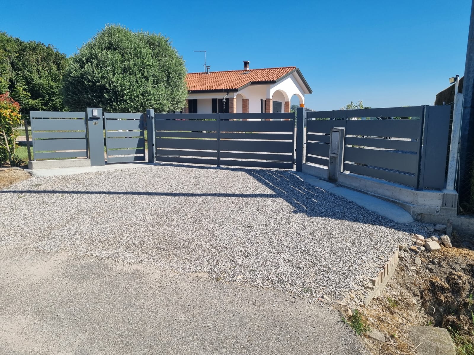 Cancello e recinzione grigi del vialetto d'accesso davanti a una casa con vialetto di ghiaia in una giornata di sole.