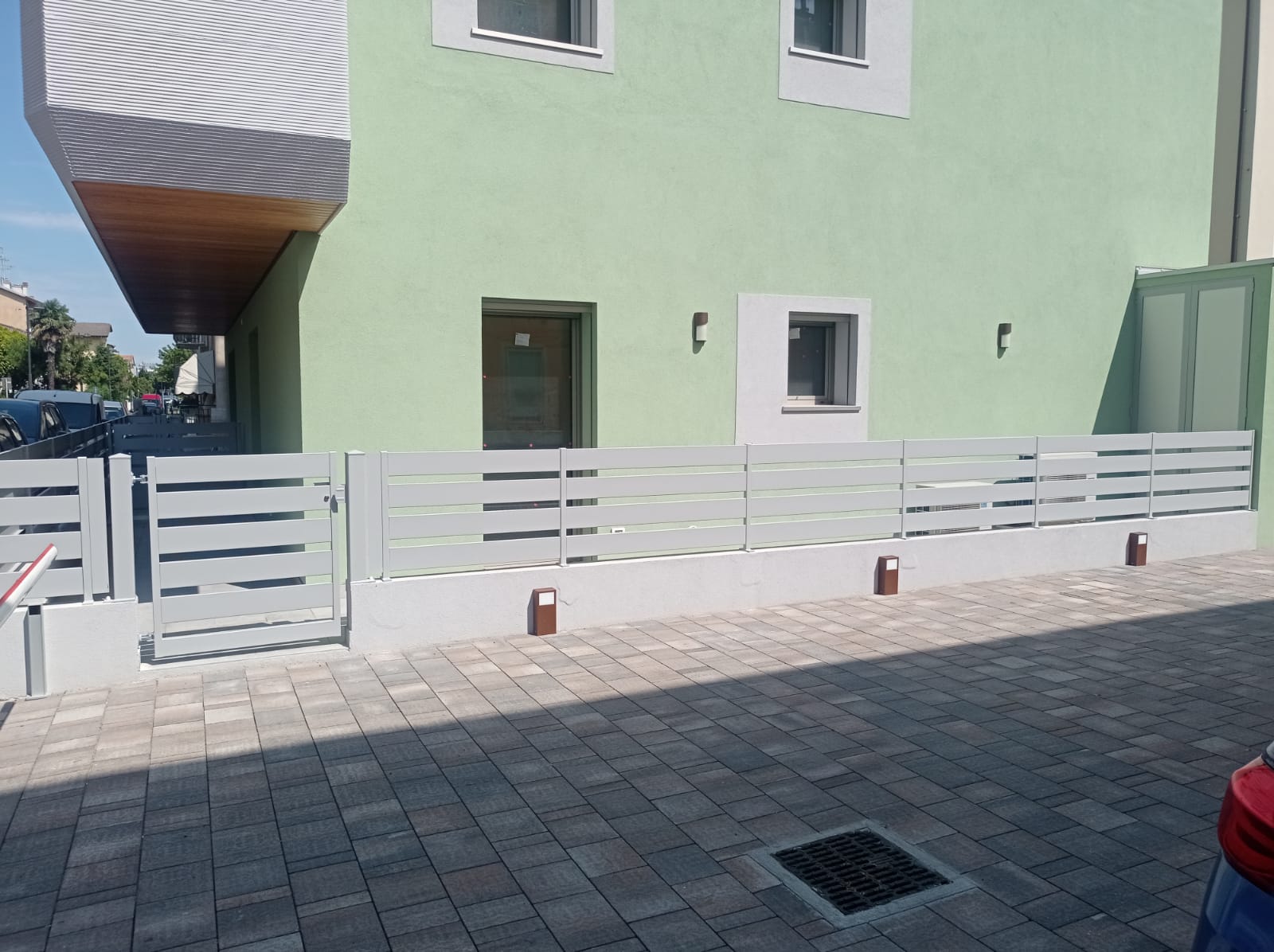 Edificio verde chiaro con recinzione orizzontale grigia sul davanti. Area pavimentata.