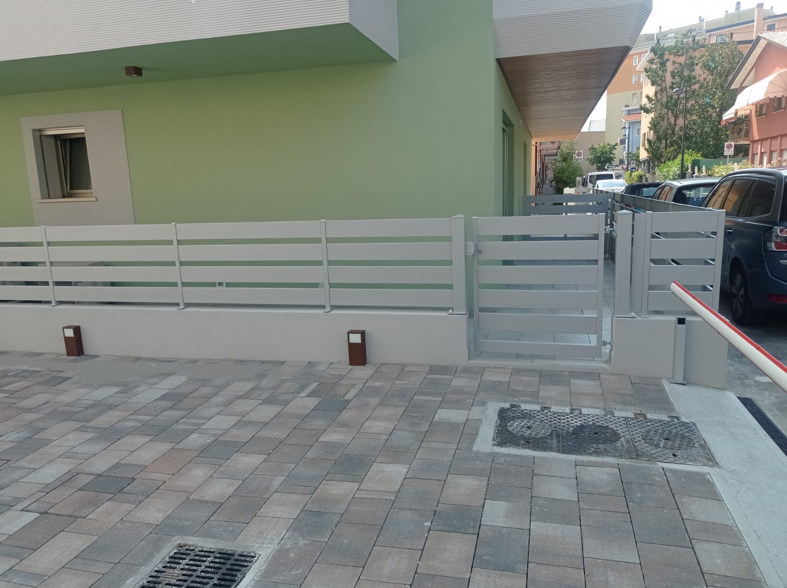 Vista esterna di un edificio con recinzione e cancello grigio chiaro, situato su un vialetto lastricato in mattoni.