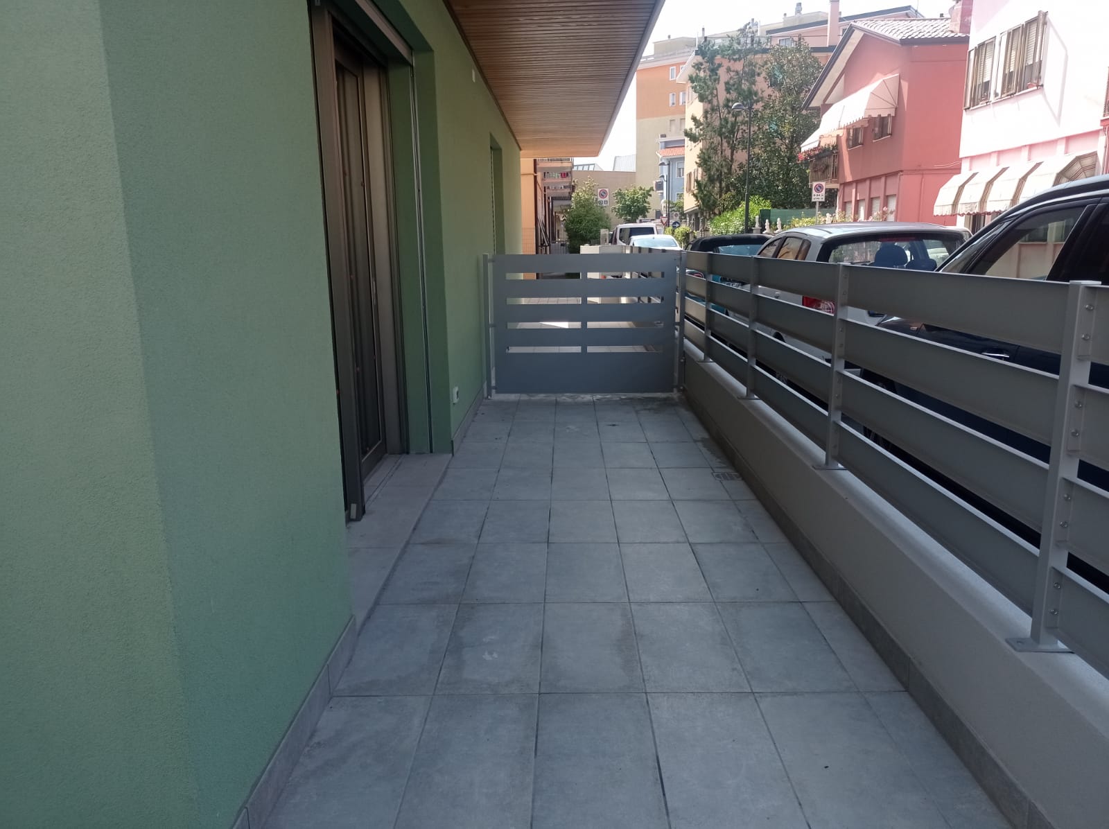 Uno stretto balcone con piastrelle grigie, un cancello di metallo e una ringhiera bassa accanto a un edificio verde.