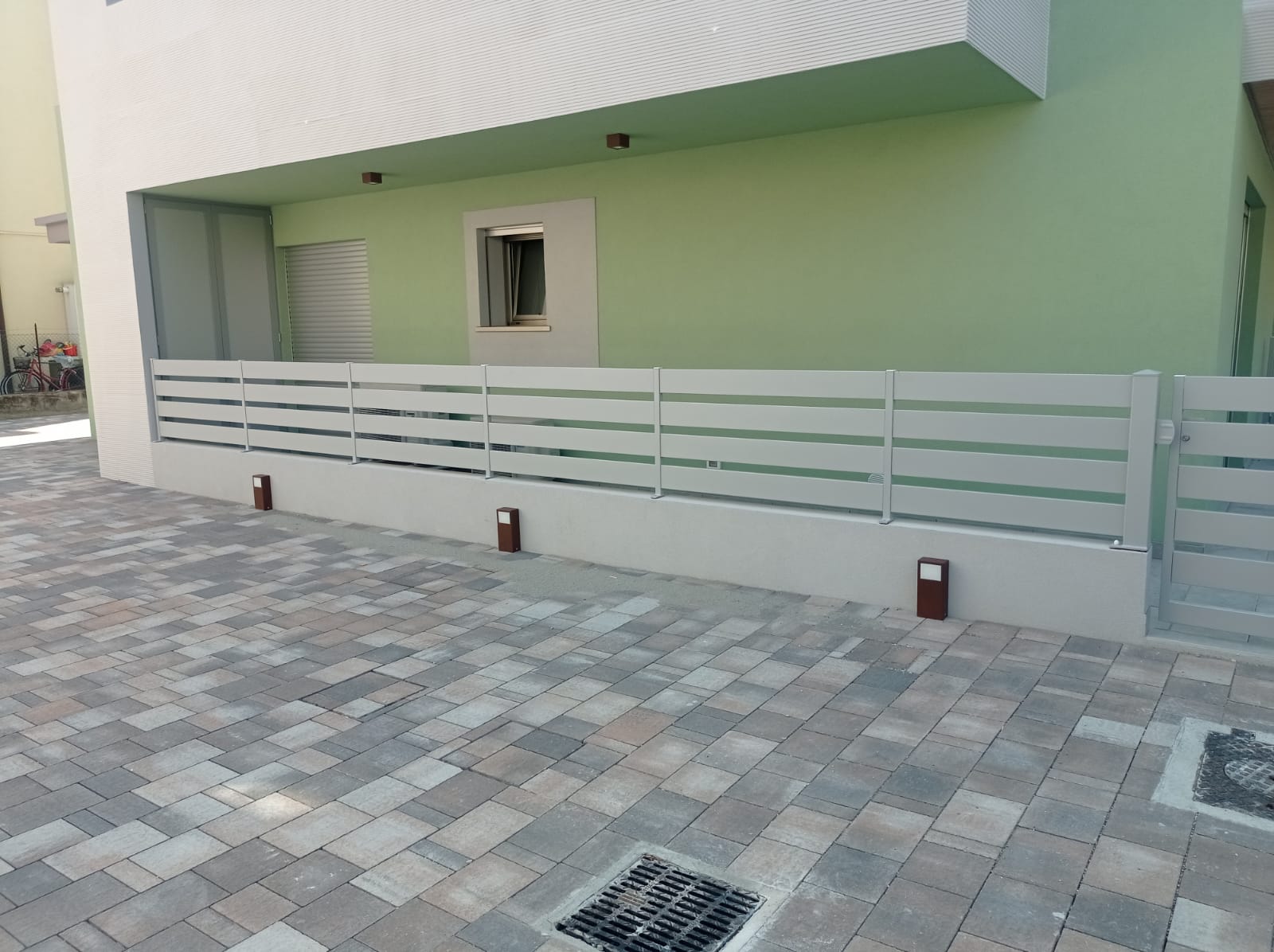 Recinzione a listelli orizzontali grigio chiaro davanti a un edificio verde chiaro. Terreno asfaltato, luci rosse.