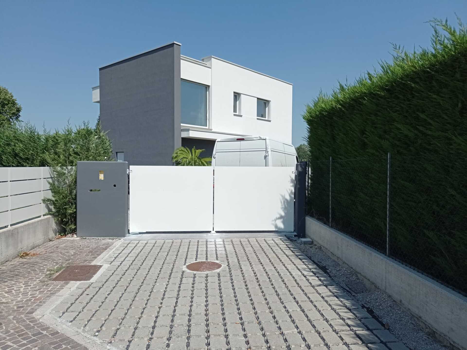 Casa bianca con vialetto d'accesso e cancello scorrevole bianco, siepi verdi, pavimentazione grigia.