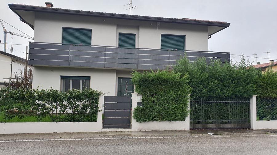 Casa a due piani con persiane verdi e balcone. Una siepe verde davanti alla casa e un cancello.